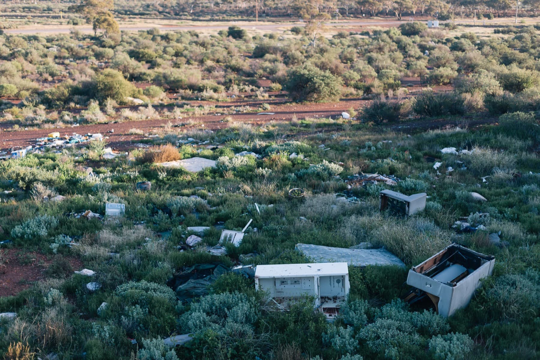 A bushland dumping ground at dawn: fridges, mattresses, a couch