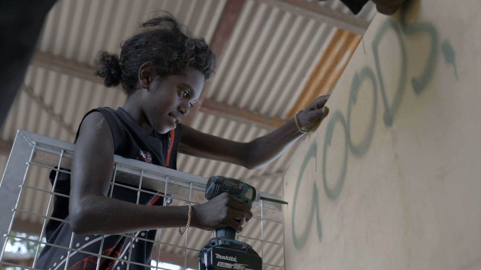 A young person drilling beside the Goods wordmark sprayed on a wall at Gamardi