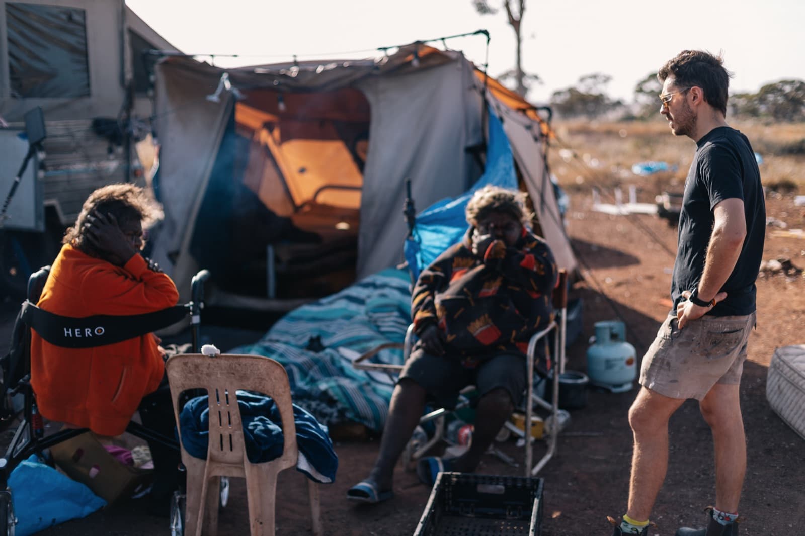 Two Elders sitting at a camp beside a tent with bedding on the ground, and Nic Marchesi standing, listening