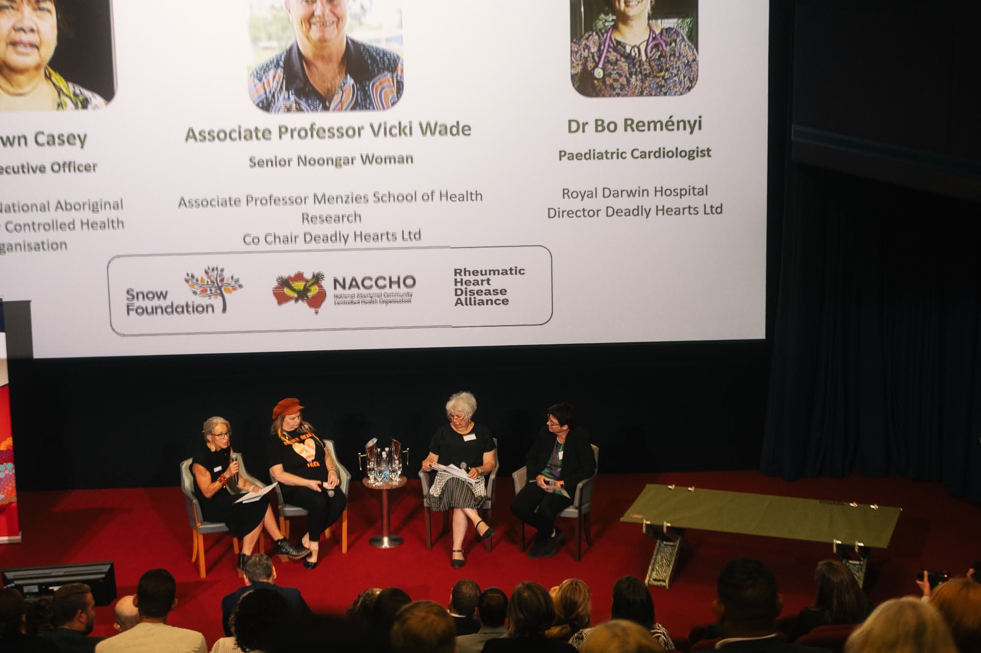 A panel on stage at Parliament House with Snow Foundation, NACCHO and RHD Alliance logos behind them, and a Goods Stretch Bed on the stage