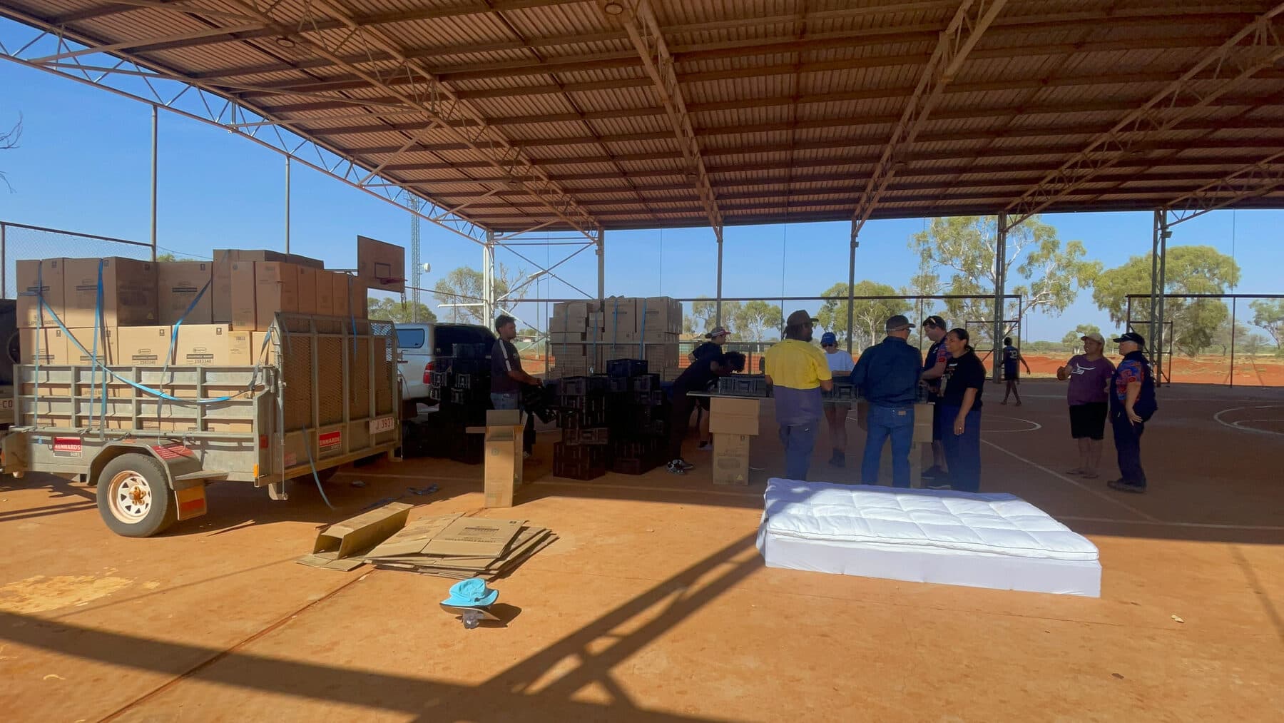 Boxes, bed parts and a finished mattress staged under a shaded court at Utopia Homelands